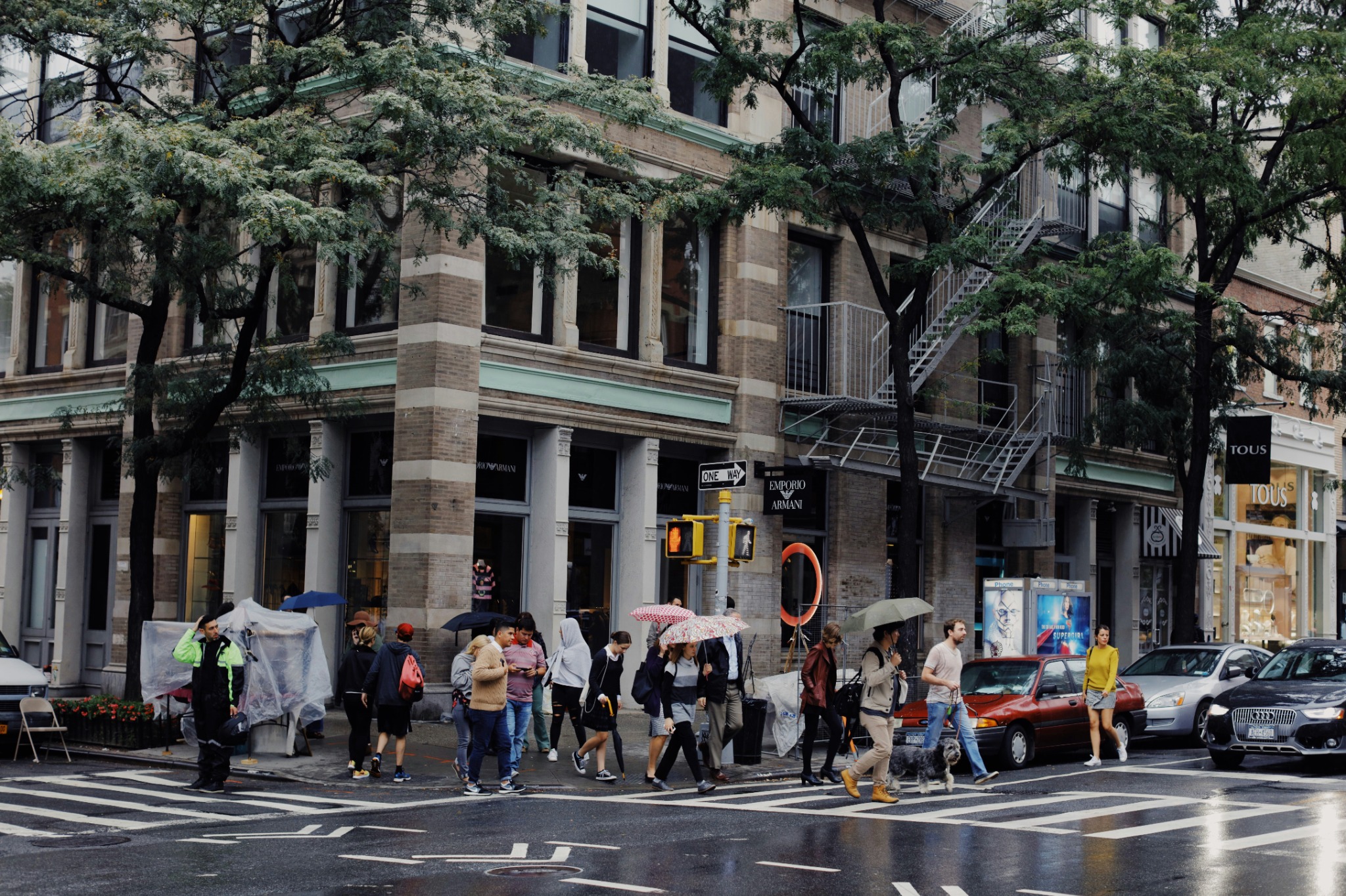 Pedestrians with umbrellas cross a rain-soaked New York street beside leafy trees and a building with fire escapes.