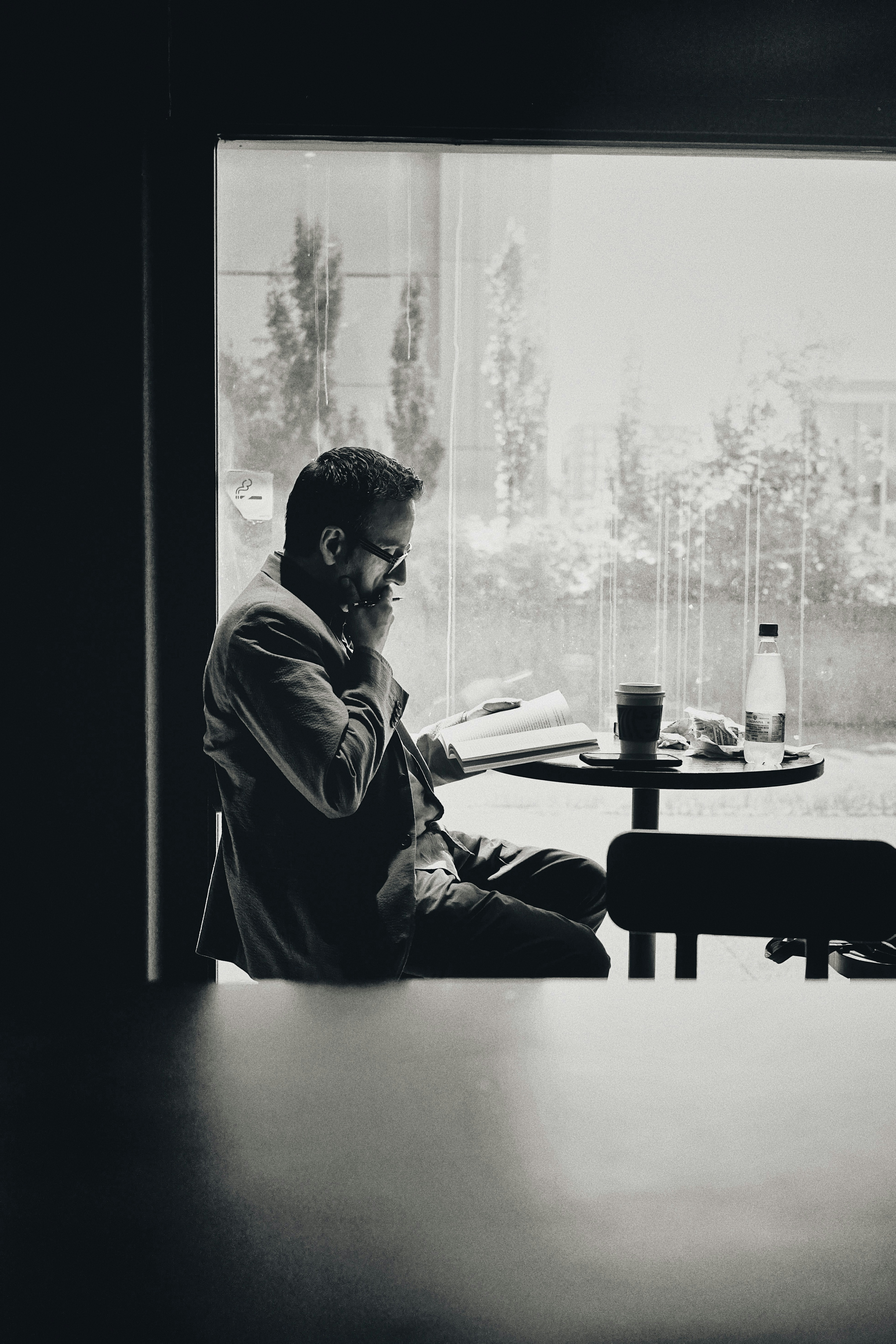 A man in glasses and a jacket reading a book at a café window, photographed in black and white.