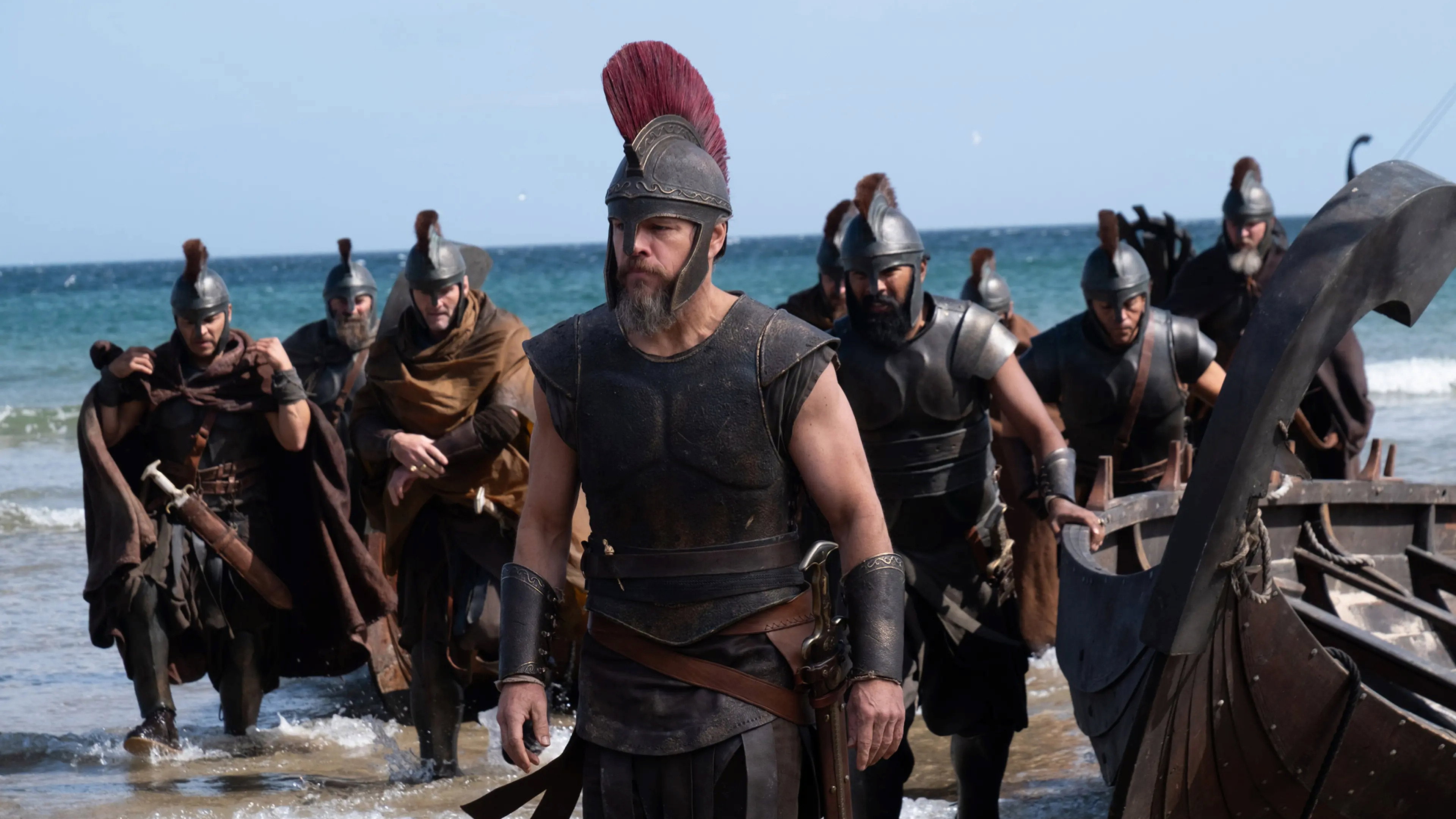Matt Damon as Odysseus in armor, coming ashore with his men beside a ship in The Odyssey.
